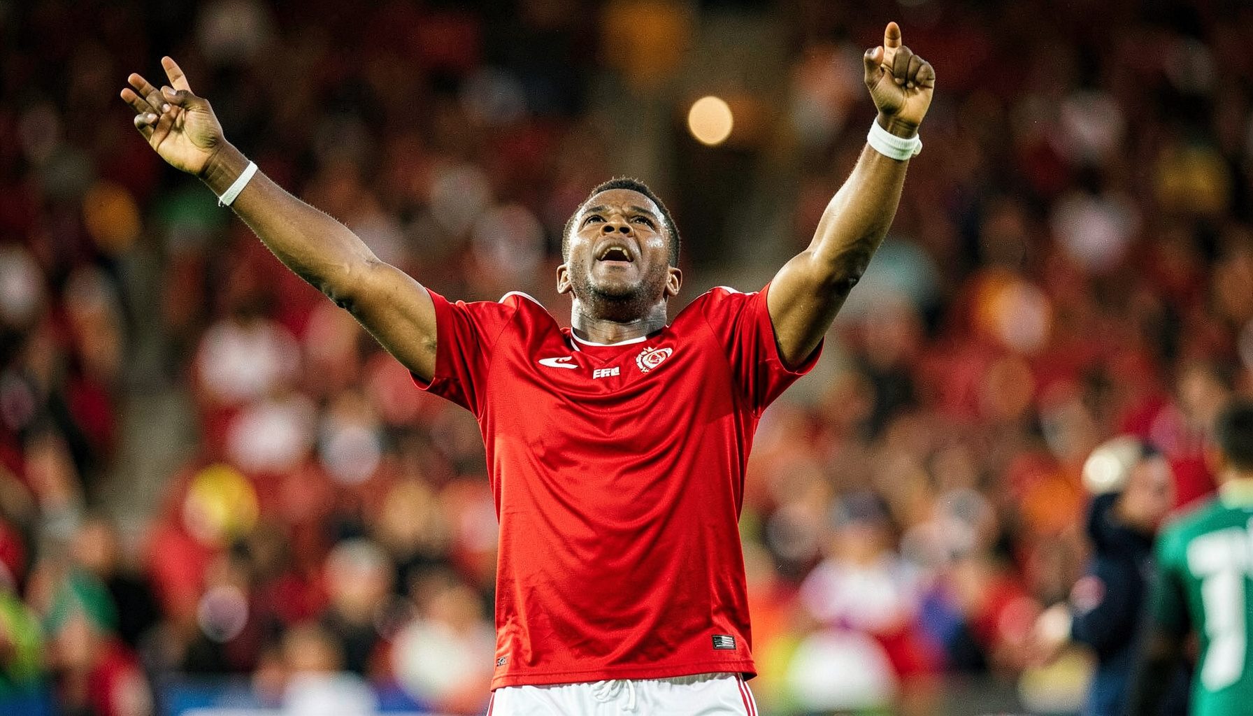 A footballer in a red jersey celebrating on the pitch, arms raised, stadium crowd blurred behind in warm floodlight glow.