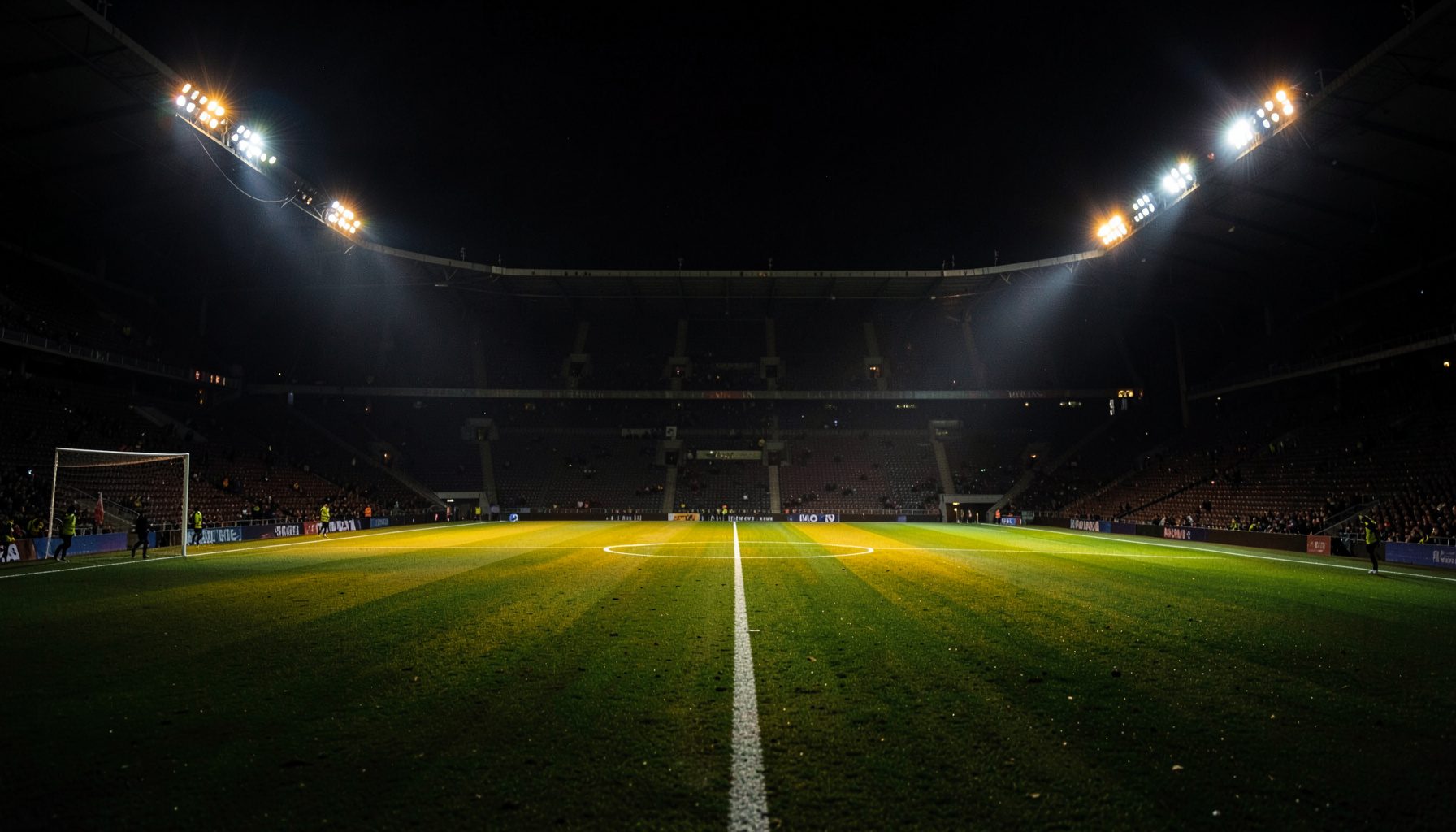 A dramatic football stadium at night under floodlights, deep green pitch with amber and white light spilling across the field, dark stands receding into shadow.